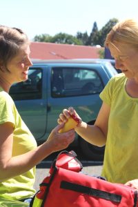 Dagmar Schmidt und Yvonne Ziegler stehend; vor Ihnen rote Tasche mit Pflegeuntensilien; Yvonne Ziegler überreicht Dagmar Schmidt eine gelben Behälter mit rotem Deckel.