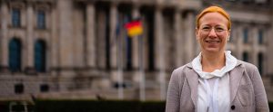 Eine Fotografie von Dagmar Schmidt, die vor der Hauptseite des Reichstagsgebäudes steht. Sie ist von der Brust aufwärts fotografiert und lächelt freundlich. Ihr langes rotes Haar ist zu einem Zopf gebunden und sie trägt einen beigen Blazer, darunter ein weißes Oberteil, sowie ihre runde rosegoldene Brille. Der Hintergrund ist unscharf. Man sieht eine schwarz-rot-goldene Deutschlandflagge und die helle, mit Säulen und großen Fenstern geschmückte, Fassade des Reichstagsgebäudes.