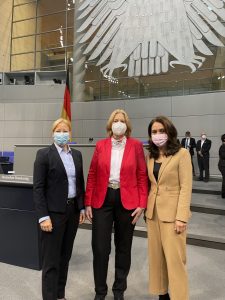 Eine Fotografie von Dagmar Schmidt (links), Bärbel Bas (mitte) und Aydan Özoğuz (rechts) im Plenarsaal des Bundestags. Die drei Frauen stehen nebeneinander und sind vom Bauch aufwärts fotografiert. Dagmar Schmidt (MdB) trägt einen schwarzen Blazer, darunter eine hellblaue Bluse, sowie ihre rosegoldene runde Brille. Ihr langes rotes Haar ist zu einem Zopf gebunden. Bärbel Bas (Präsidentin des Bundestags) trägt einen roten Blazer, darunter eine weiße Bluse. Ihre blonden schulterlangen Haare sind offen. Aydan Özoğuz (Vize-Präsidentin) trägt einen braunen Blazer, darunter ein schwarzes Oberteil und ihr braunen schulterlangen Haare ebenfalls offen. Alle Drei tragen einen medizinischen Mundschutz. Hinter ihnen ist ein Ausschnitt des Plenarsaals zu sehen. Links stehen ein paar der charakteristischen blauen Bürostühle. Mittig im Hintergrund ist das Podest zu sehen, auf dem Präsidentin und Stellvertreter:innen Platz nehmen. Davor stehen das graue Rednerpult mit dem Schriftzug „Deutscher Bundestag“ und dahinter eine große Deutschlandflagge. Am oberen Bildrand sind noch die Füße und Schwanzfedern des grauen metallenen Bundesadlers zu erkennen.